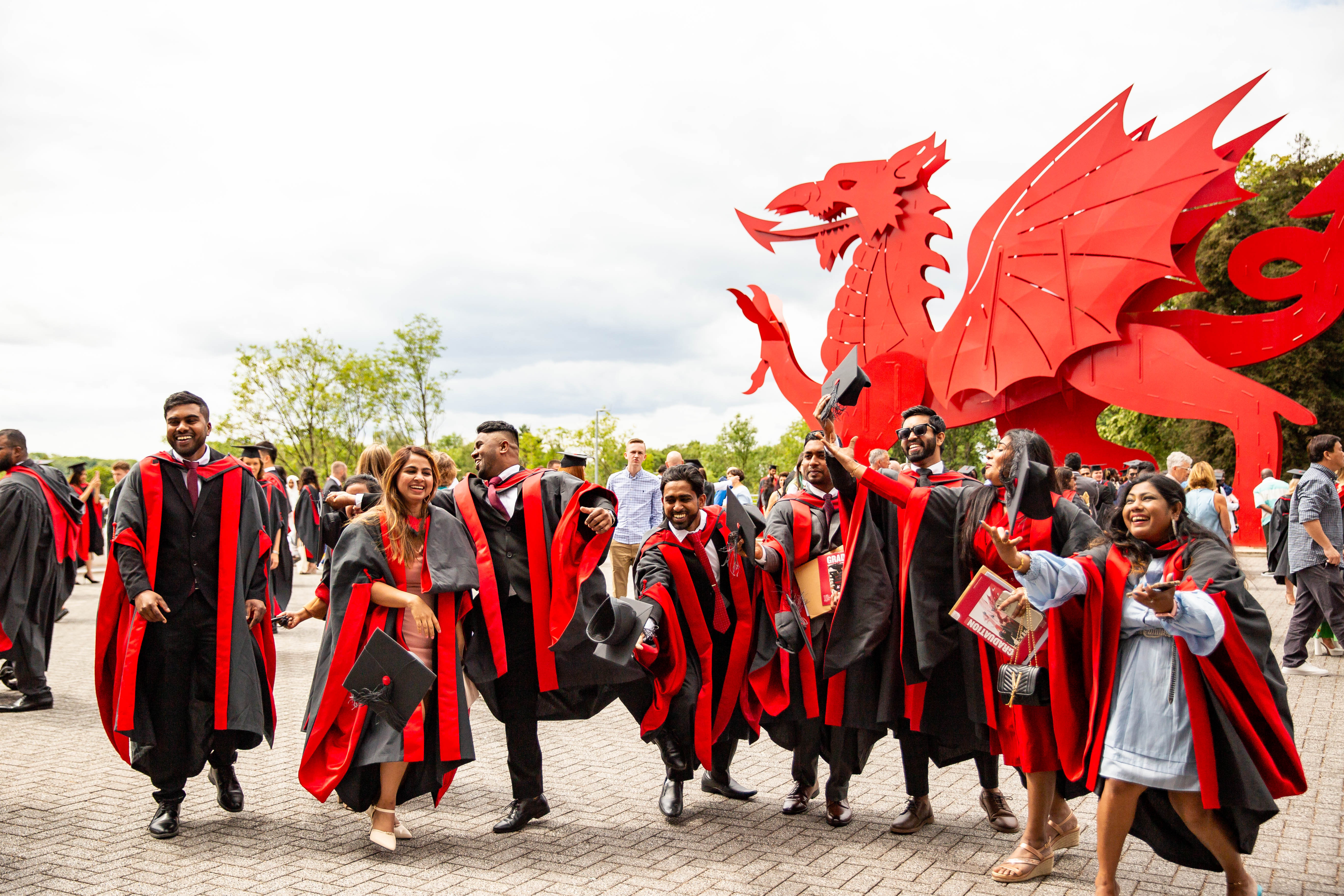 A group of students in graduation gowns throwing caps into the air in front of the Welsh dragon.