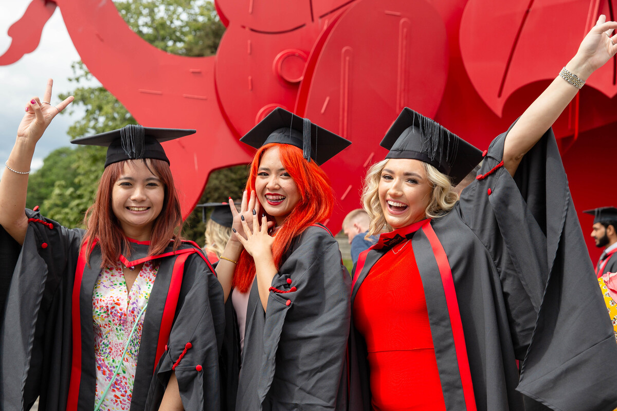Three students dressed in graduation robes and graduation caps smile and pose in front of the red dragon outside Newport's ICC
