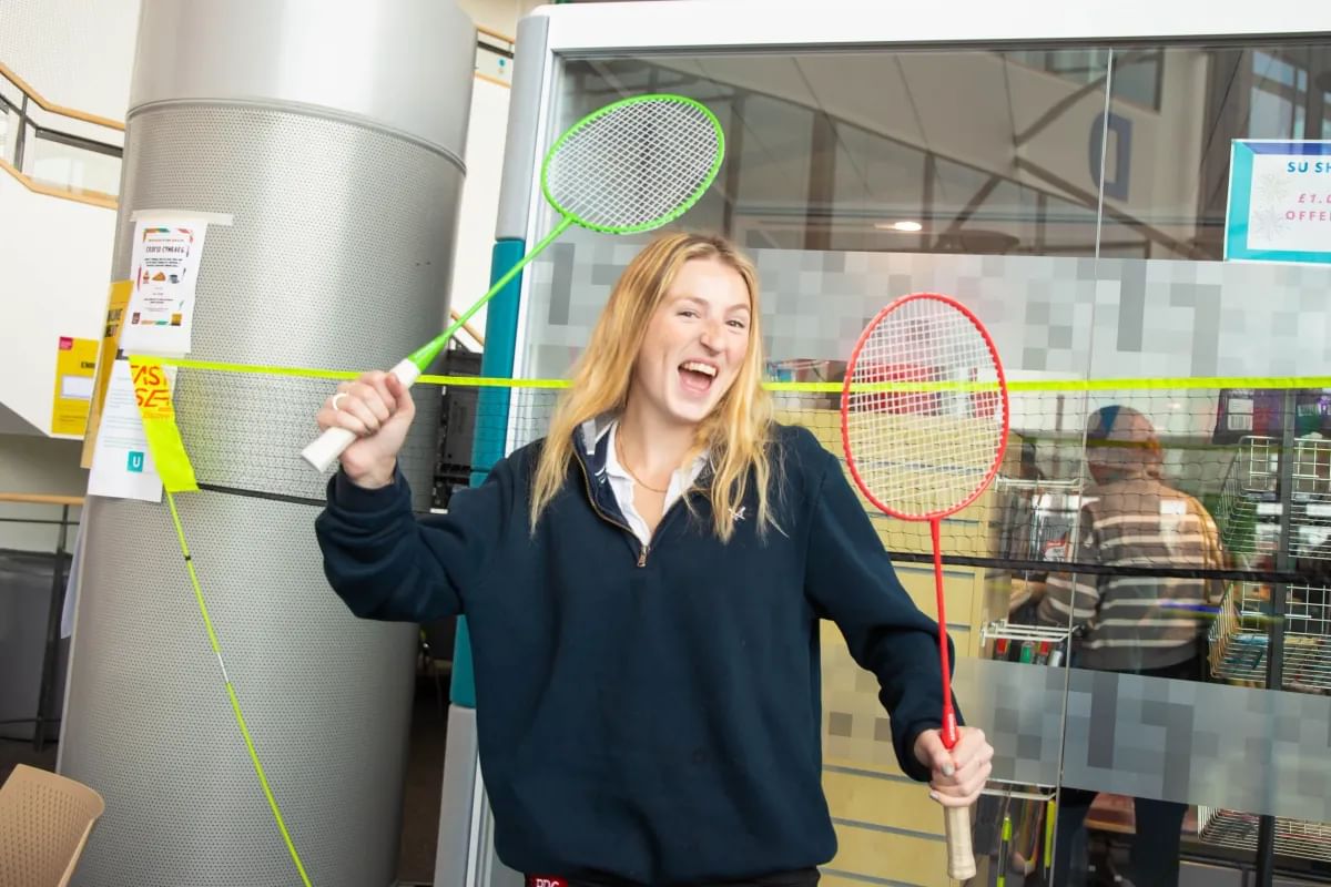 A smiling student holds two badminton rackets in front of a glass door at a Newport welcome event for students.