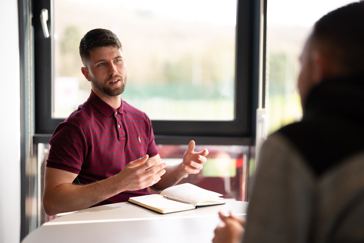 A student talking whilst sat at a table with a book in front of him.
