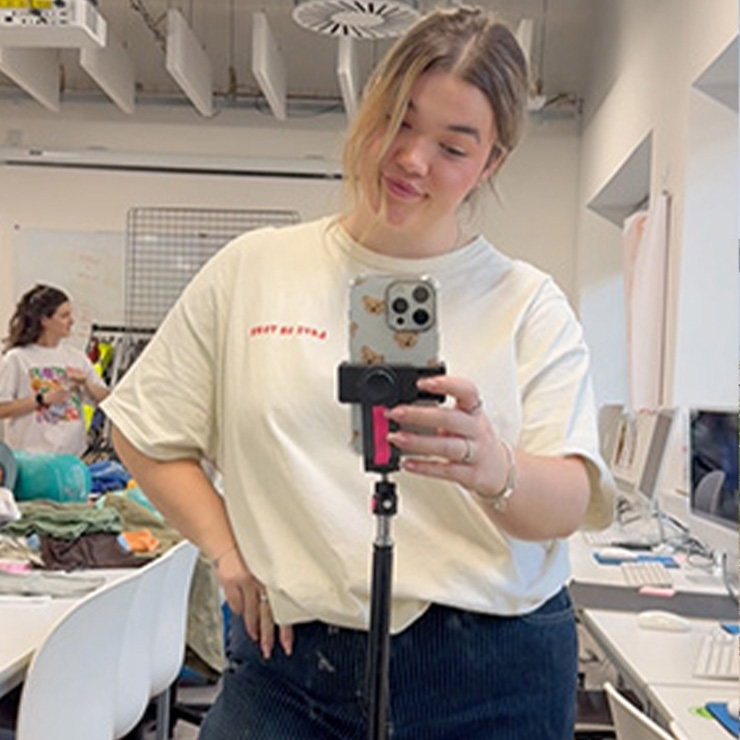 A student is standing in front of a mirror and taking a selfie on their phone, using a tripod.