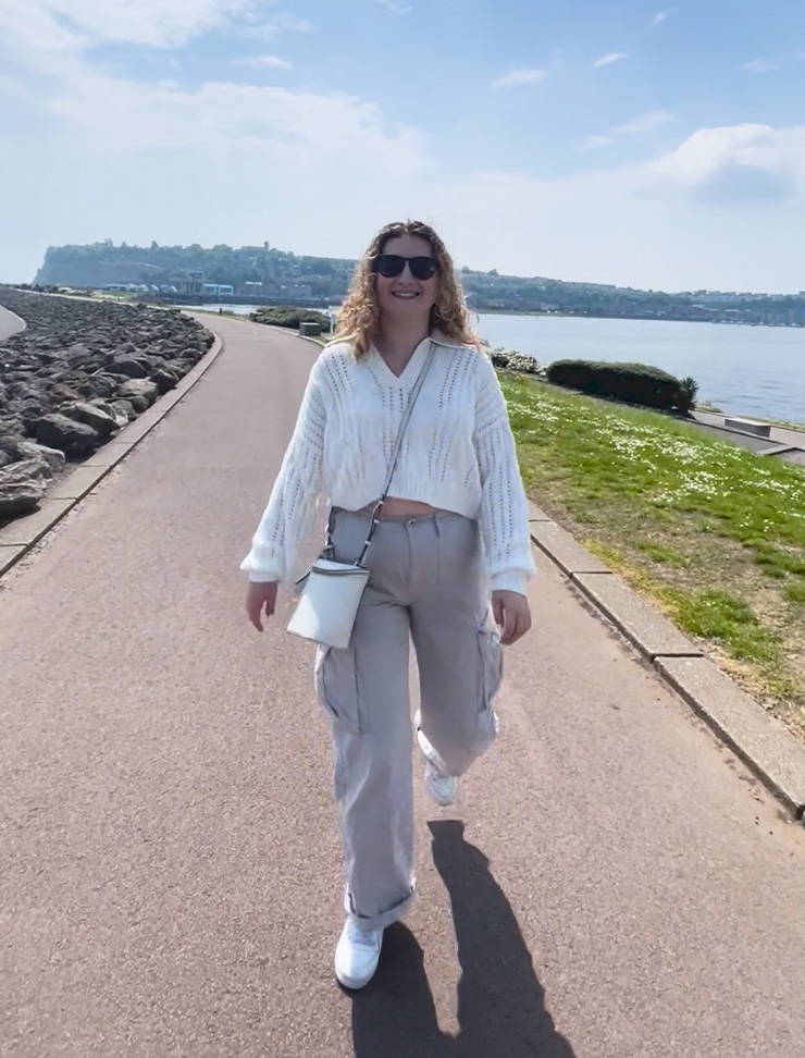 A student is walking along Cardiff Bay, smiling at the camera.