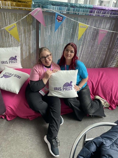 Two students sat on a bright pink seating area, holding a cushion that reads 