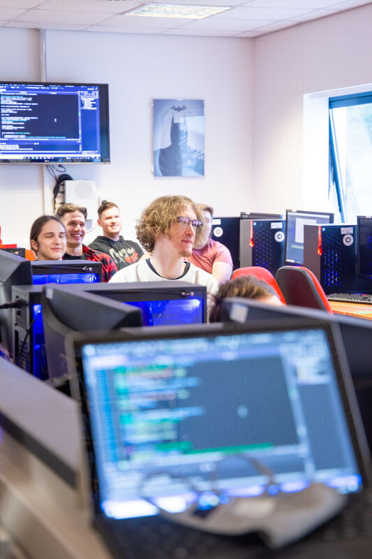 A class of computing students working in a classroom. In the foreground there is a blurred laptop screen with coding on it.