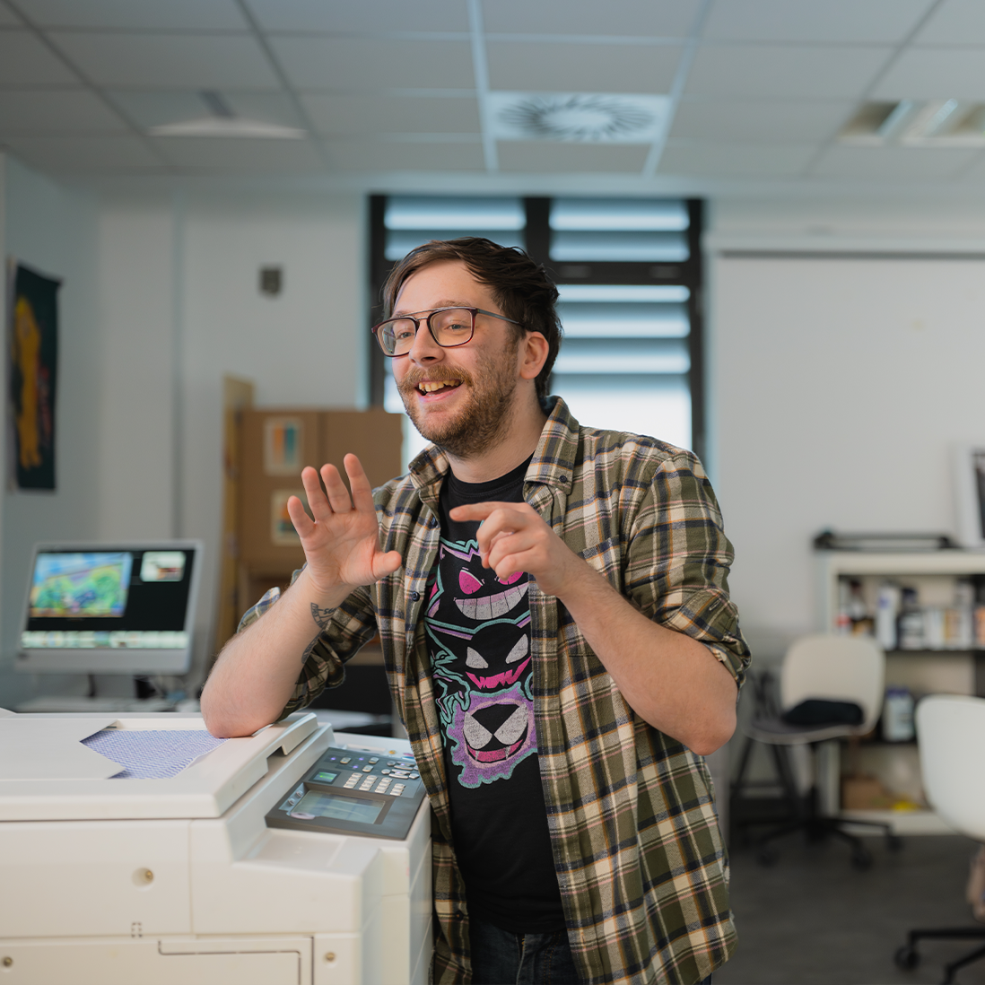 Illustration student, James, is leaning his elbow on a printer whilst talking and gesturing.