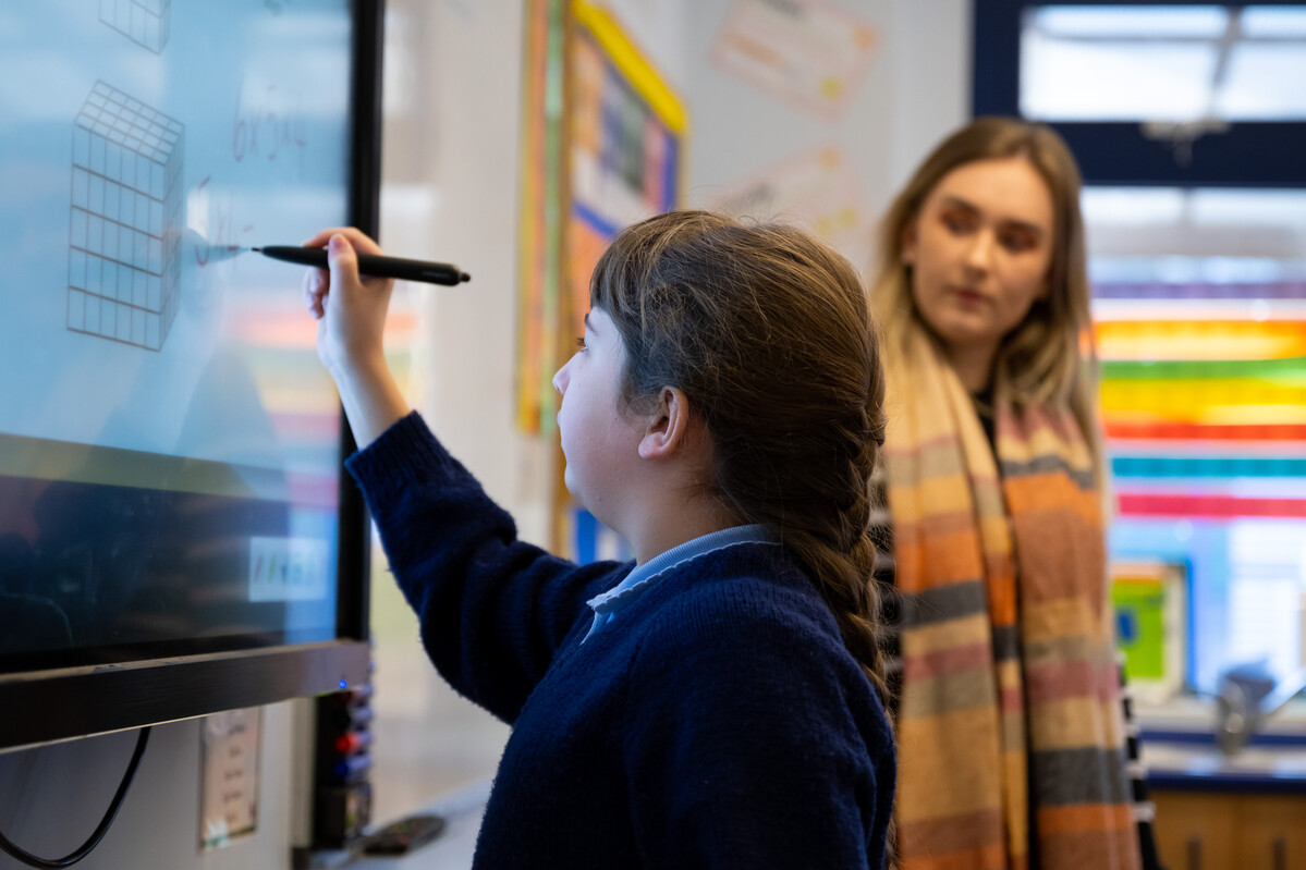 teacher looking at young pupil as she interacts with a digital whiteboard with stylus pen
