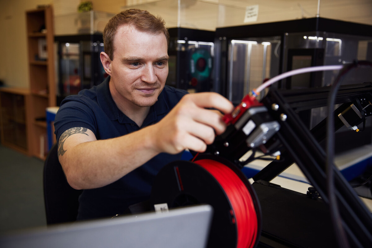 A student is altering some cable in an engineering lab, behind is a row of 3D printers.