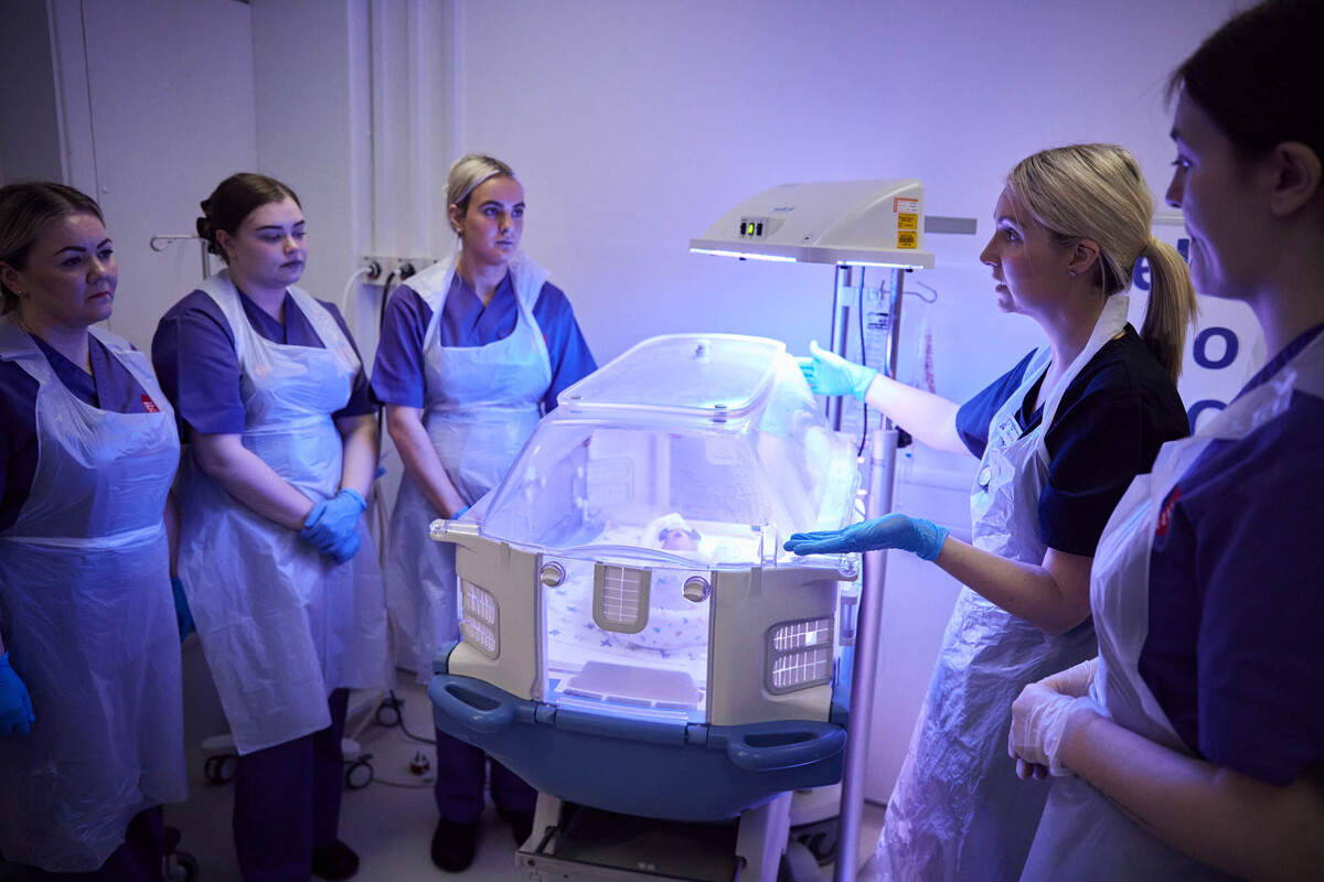 A group of Midwifery students are listening to a talk around a fake babies incubator.