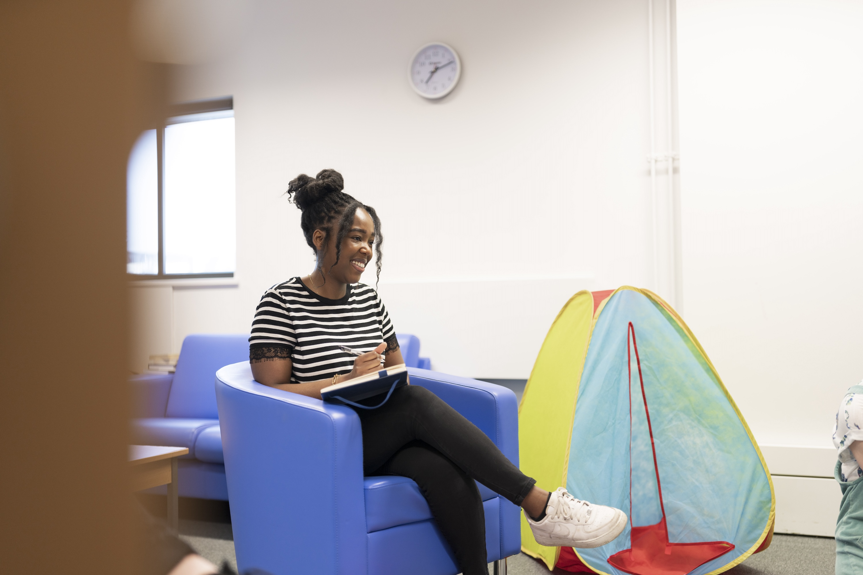 A psychotherapy and counselling student smiles as they write in their notepad while sat in a blue chair next to a child's tent in play therapy classroom