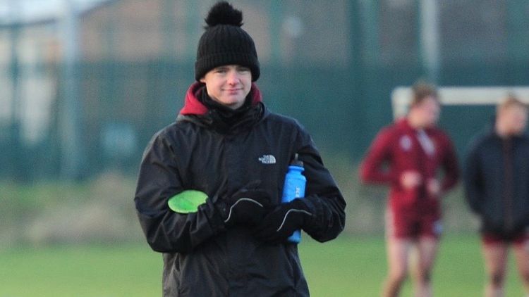 A football graduate in a black jacket and hat trains players, holding a green cone on the pitch.