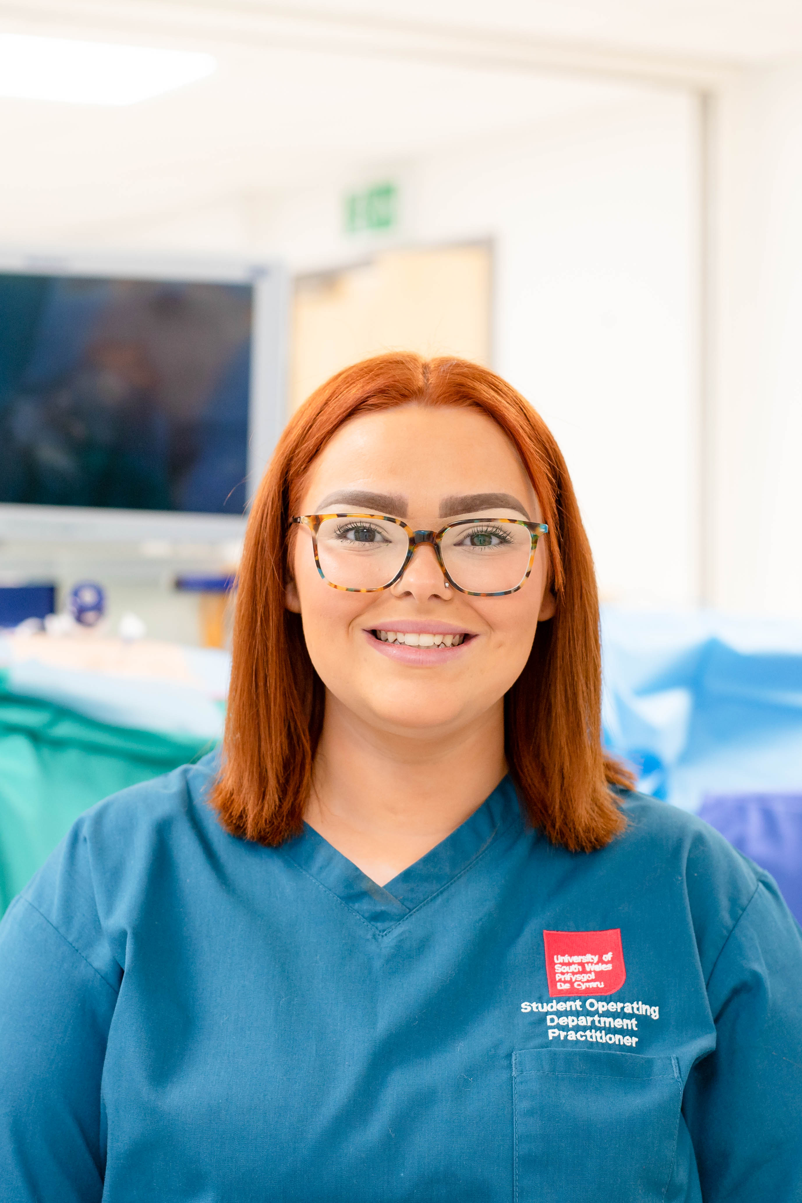 ODP student Hollie smiles directly at the camera whilst wearing her blue scrubs. There is medical equipment behind.