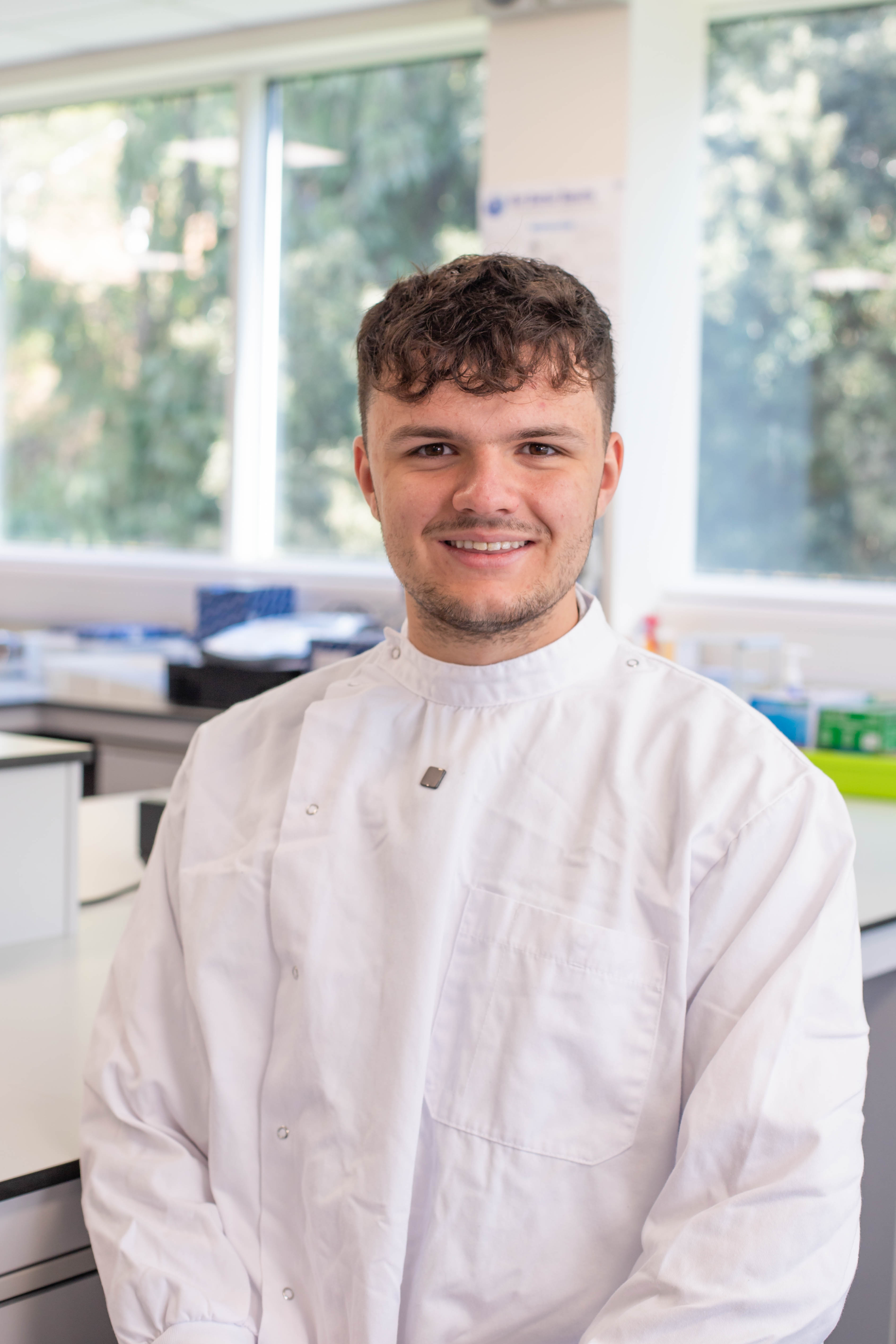 Biological Science student, Oliver, smiles for a profile shot in a science lab, wearing a white lab coat
