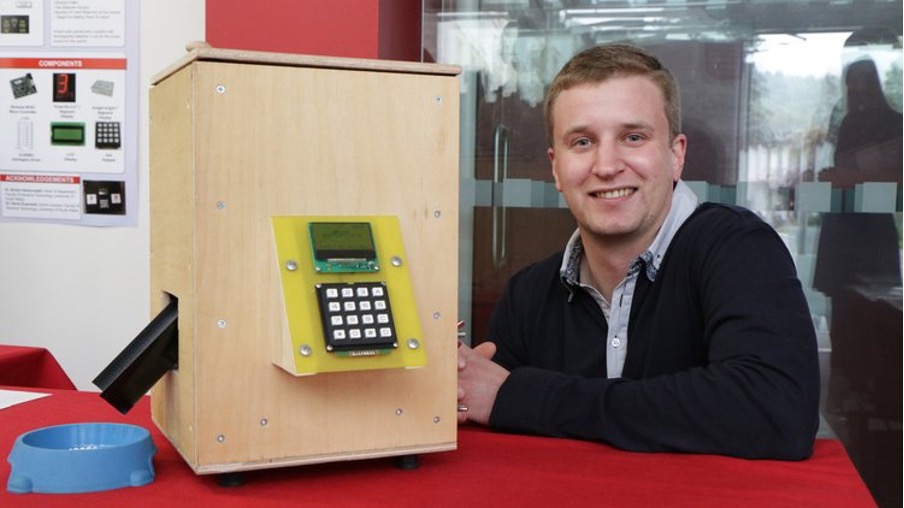 Graduate, Edwin, smiles at camera whilst displaying his cat feeder invention