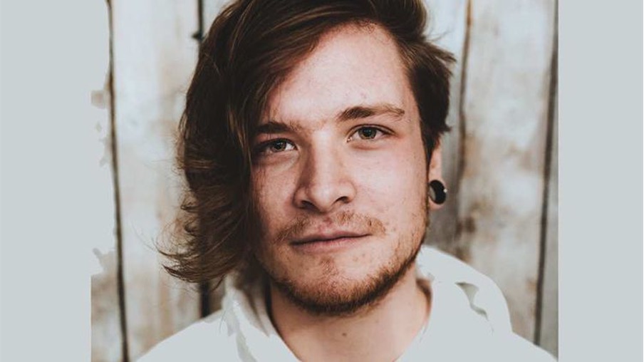Music graduate, James, looks directly at camera whilst having his photo taken against a white wooden wall