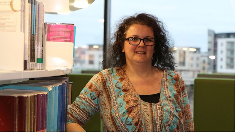 Teaching student, Julia, smiles at the camera whilst leaning on a bookshelf in Newport campus
