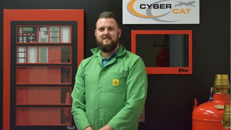 Shaun poses for a photo in his workplace. He is wearing a green zip-up coat.