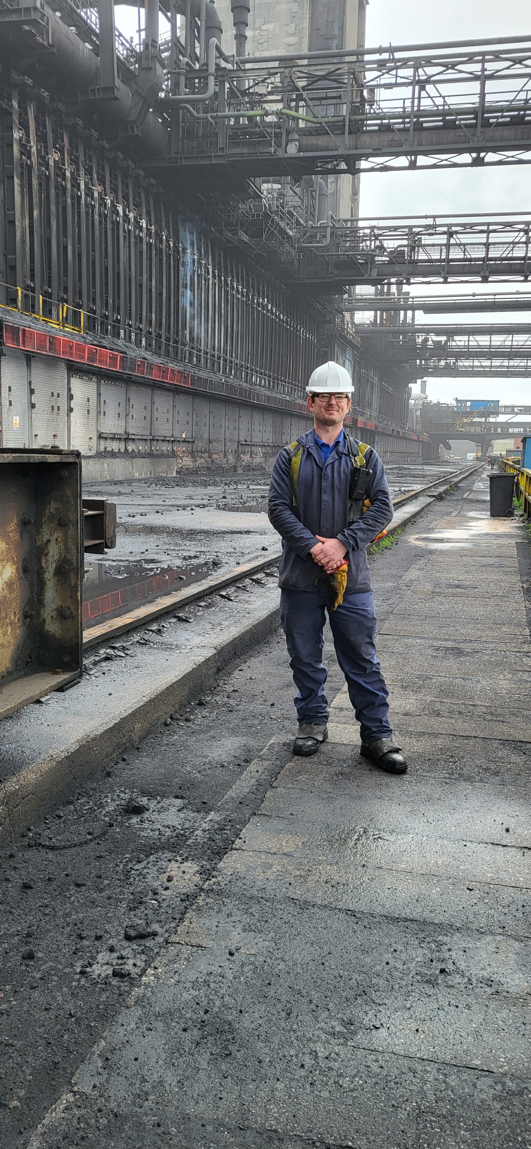 Engineering graduate Wayne stand proudly at his workplace wearing blue overalls and a hard hat