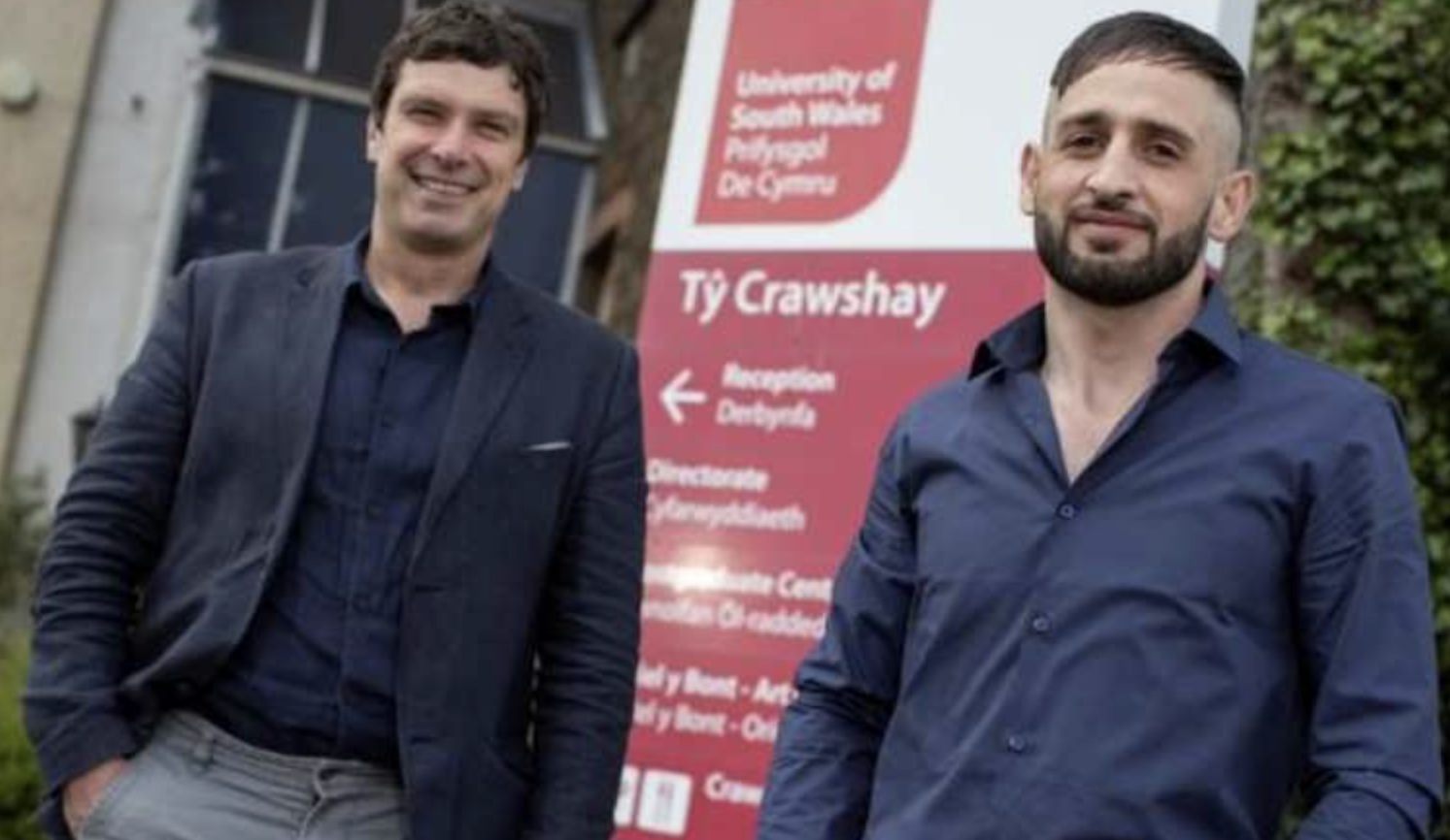 Dr Mike Chick and Tarek Zou Alghena both smile at the camera while stood together and dressed smart casual in shirts and smart jeans in front of a USW directions sign pointing to Ty Crawshays on Treforest campus