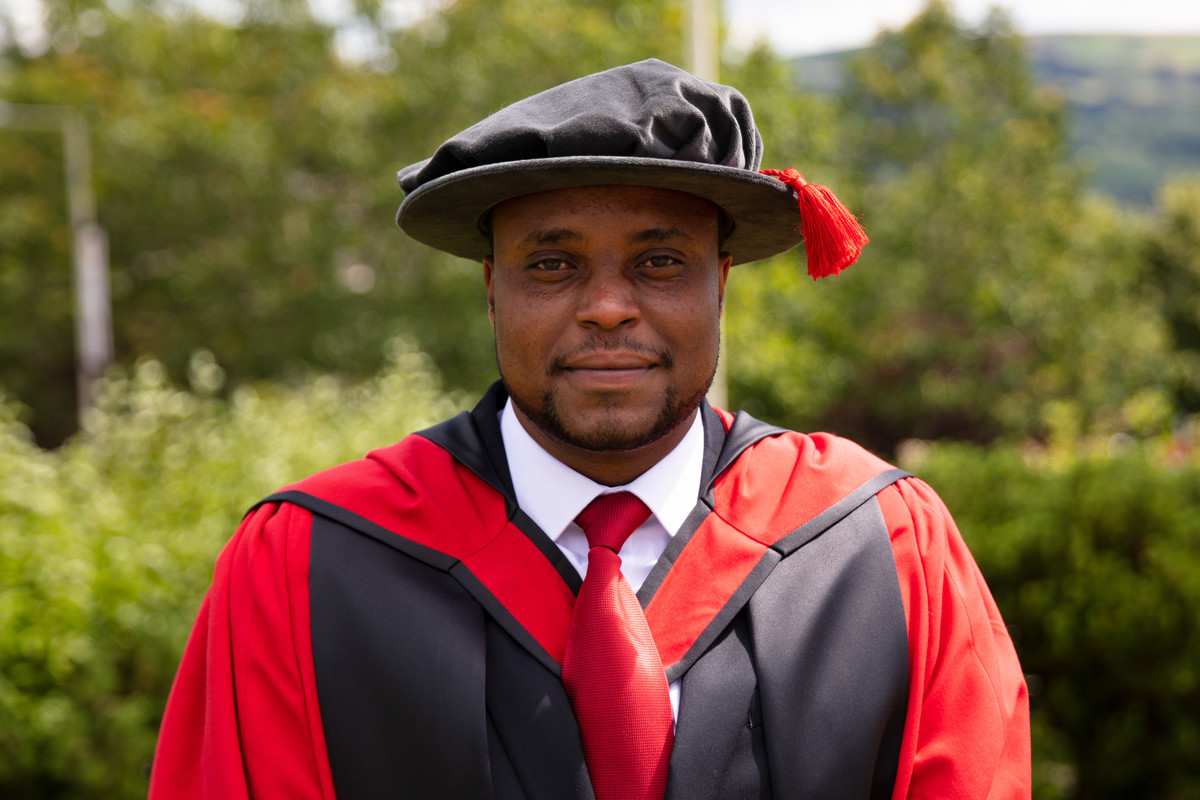 Prosper Mbowe PhD stood smiling, wearing academic robes and graduation tam.