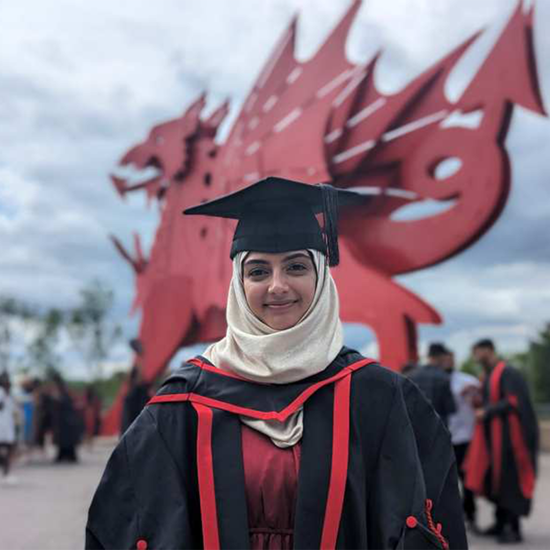 Graduate Mareah Ali wearing a graduation robe and graduation cap stood in front of the red dragon outside Newport's ICC
