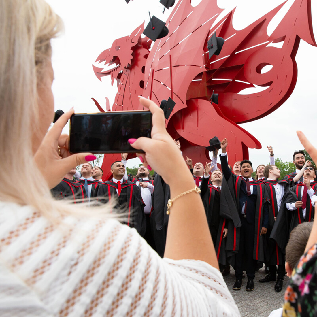 A person takes a photo on their phone of a large group of graduates, dressed in graduation robes, trying to catch their graduation caps after throwing them in the air by the red dragon outside Newport's ICC