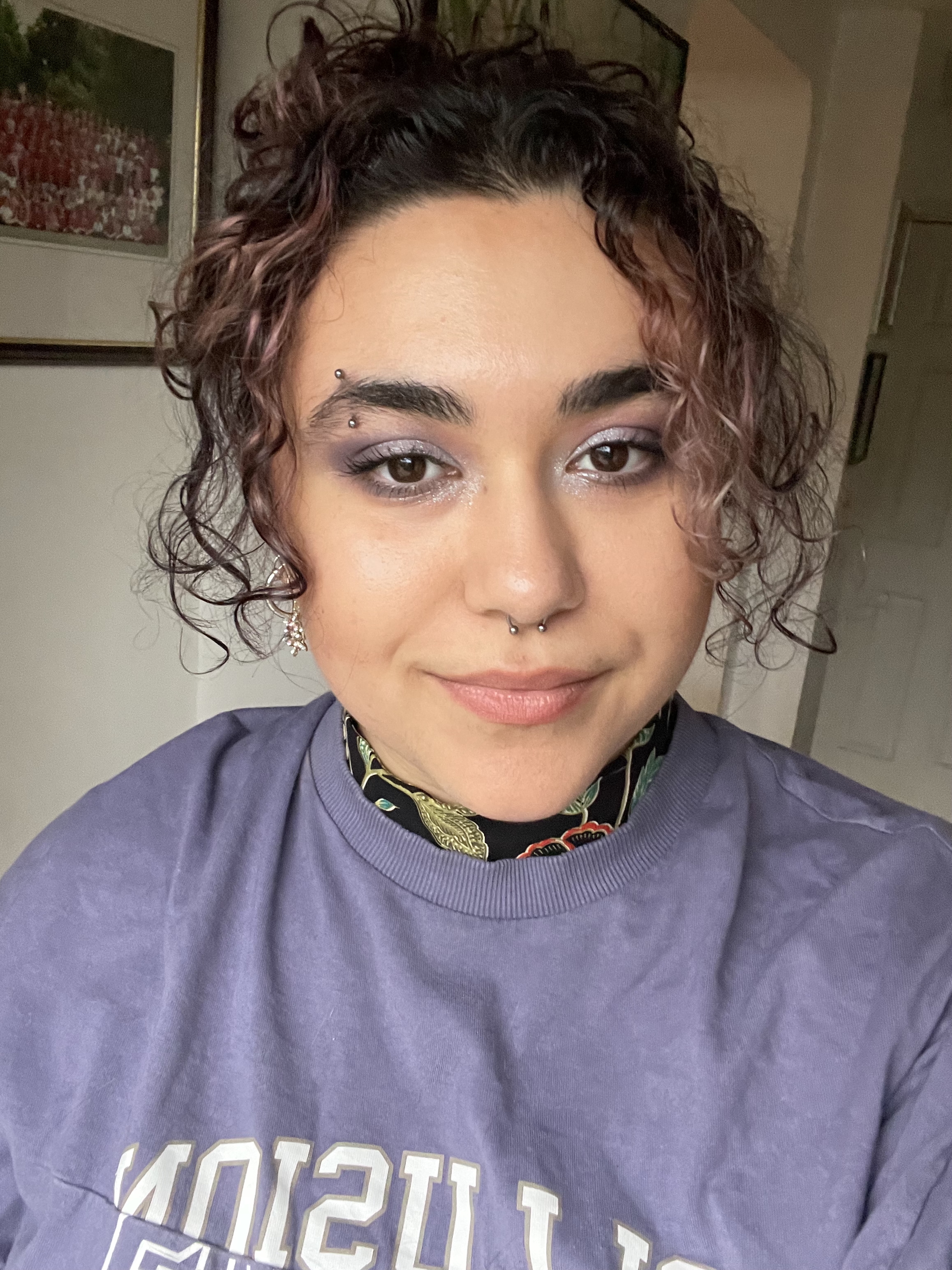 A person smiling and posing for a selfie indoors, wearing a purple shirt.