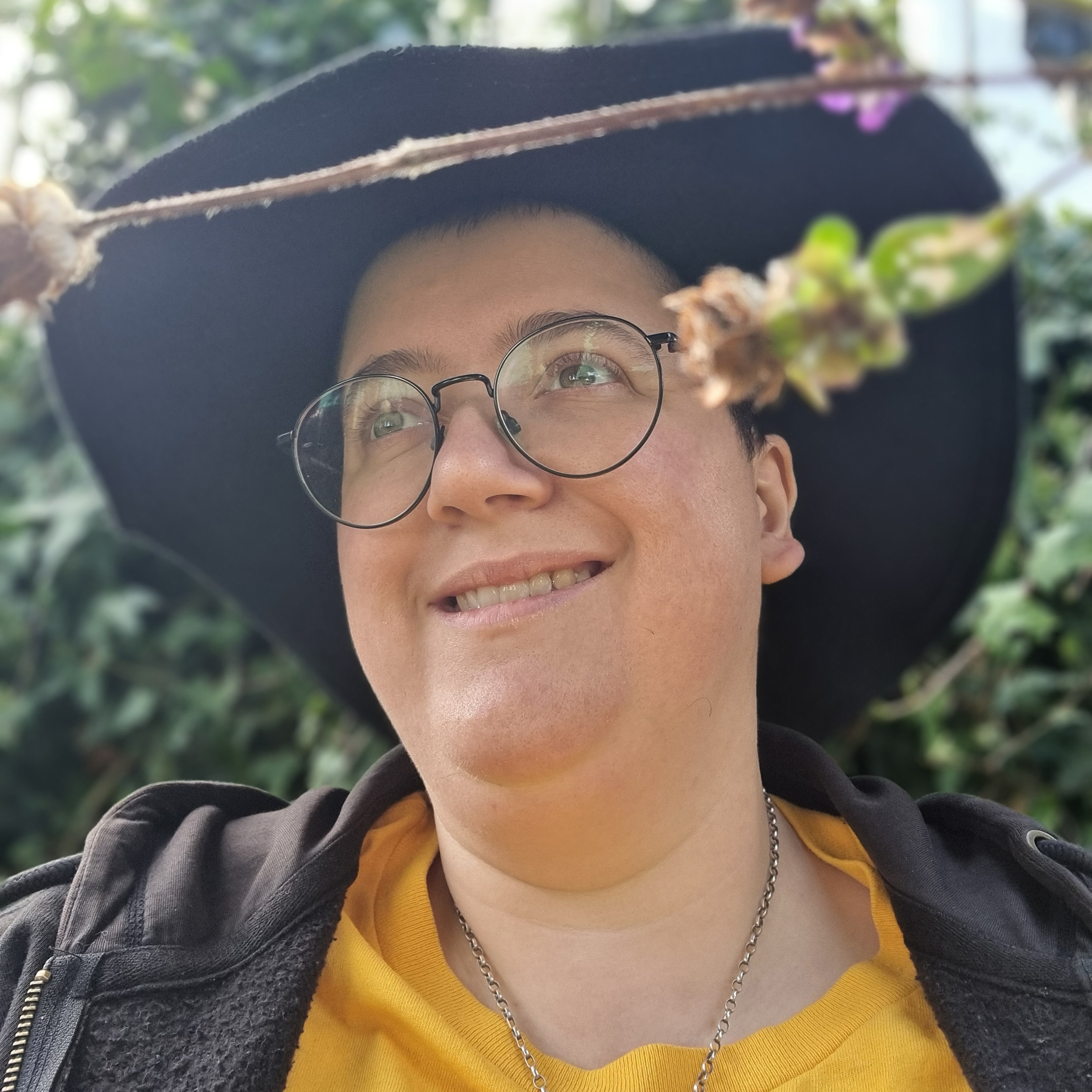 photograph of a person with a large black hat and wearing glasses. Flowers are in the foreground.